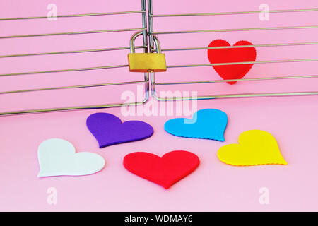 One red silk heart is in a cage locked with padlock. Five multicolored hearts are outside. To find the true love among people and loneliness concepts. Stock Photo