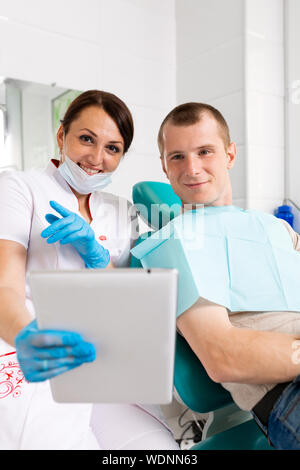 Happy dentist and patient commenting treatments in a tablet application in a consultation with medical equipment in the background. Stock Photo