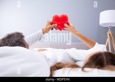 Couple Lying On Bed With White Blanket Holding Red Heart In Their Hands Stock Photo