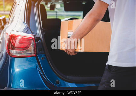 Delivery man is delivering cardboard box to customers via private car trunk door. People lifestyles and business occupation concept. Young male courie Stock Photo