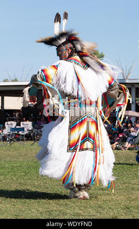 Young Native American man dancing in a pow wow in a white fringed garment with ribbons and geometric designs and feather and fur headdress. Stock Photo