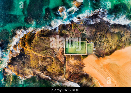 Rectangular swimming rock pool on Mona Vale beach sandstone plateau on a surf line of Pacific ocean waves in aerial top down view in soft morning ligh Stock Photo