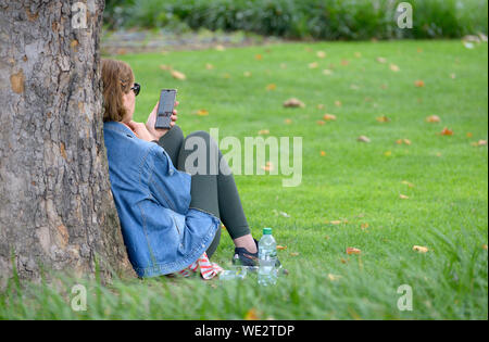 London, England, UK. Woman sitting against a tree, looking at Google on her mobile phone Stock Photo