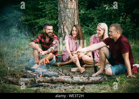 Young couples having picnic in woods. Bearded man and his best friend frying sausages over fire. Happy people sitting around campfire, friendship and Stock Photo