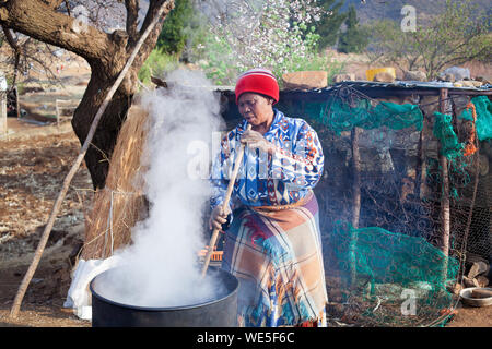 Ramabanta, Lesotho - September 20, 2017: african woman in traditional blanket clothes brews national drink in metal barrel, village courtyard, Africa Stock Photo