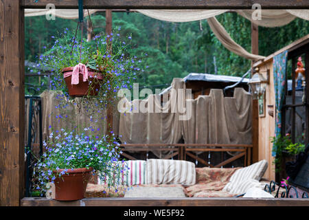 Beautiful scenery of the rustic veranda: pots with purple flowers, a lamp, decoration with various fabrics and a sofa Stock Photo