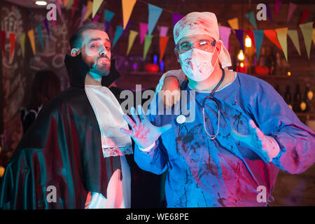 Scary man in a doctor costume covered in blood with his handsome dracula friend celebrating halloween. Halloween costumes. Stock Photo