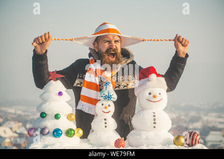 Holidays celebration concept. Angry man holding hat strings in hands. Christmas and new year. Snowmen and snow xmas tree on blue sky. Hipster shouting Stock Photo