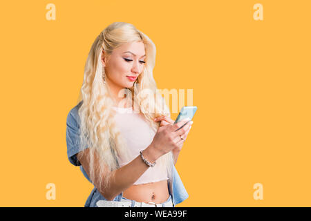 Lovely caucasian serious student with long hair, looking at screen of cell phone, making video call, reading news, using wireless internet on yellow b Stock Photo