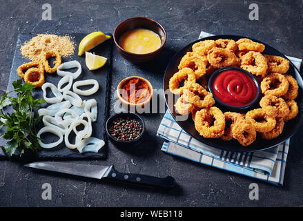 crispy calamari rings, deep fried breaded squid rings served with tomato sauce on a black plate on a concrete table with ingredients, horizontal view Stock Photo