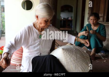 (190830) -- HANOI, Aug. 30, 2019 (Xinhua) -- Local women make traditional conical hats in the Chuong village on the outskirts of Hanoi, capital of Vietnam on Aug. 29, 2019. Vietnam's traditional conical hats are mostly made from bamboo and palm leaves, and they are among the country's most impressive traditional costumes. People in the Chuong village have been making conical hats for about 400 years. The locals buy green palm leaves from other provinces, dry them in the sun and iron and bleach the leaves into flat slices. They then use thin strings to fix layers of leaves on a cone made of bam Stock Photo