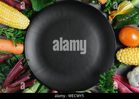 Assorted fresh vegetables on black background. Corn, tomatoes, zucchini, carrot, beetroot, garlic, bean, black plate. Health lifestyle and food concep Stock Photo