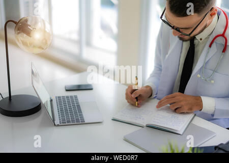 Doctor working with laptop computer and writing on paperwork with physician's stethoscope on desk in the hospital. Stock Photo