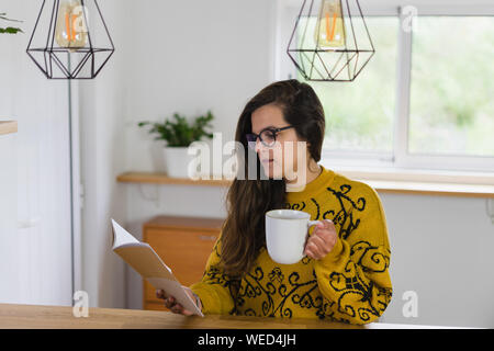 Woman with glasses holding a mug and reading from a notebook on a wooden workbench inside a house. Stock Photo