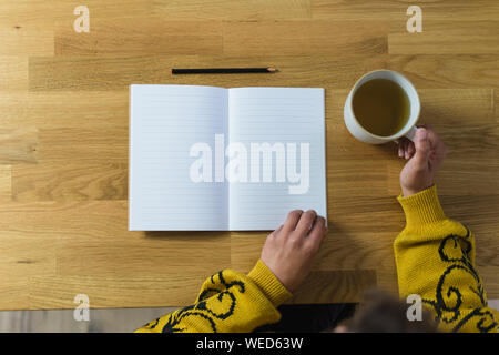 Top view of a opened notebook and a woman's hands holding a mug and the notebook in the flip page position. Wooden countertop as background. Stock Photo