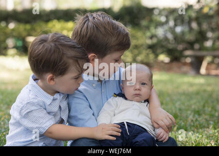 Two boys, 4 year old and 2 year old, holding their baby brother in a park Stock Photo