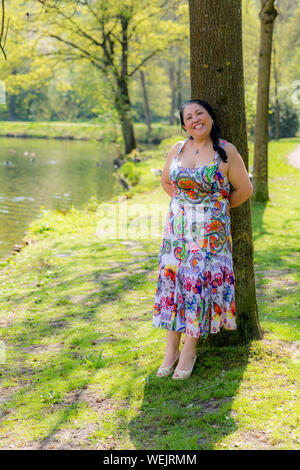 Smiling Mexican woman leaning on a tree trunk in a white dress with multicolored decorative, wonderful sunny day in the park Stock Photo