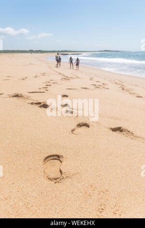 Footprints along the sand coast, a group of four people are hanging out in the beach, the ocean has just a single wave, in the horizon there are some Stock Photo