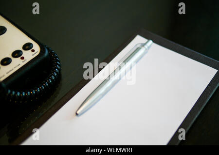 the silver pen color is placed on white short note paper on the brown wooden table near telephone in the luxury hotel room Stock Photo