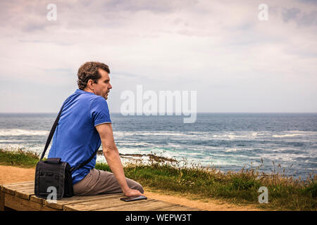 man with blue shirt sitting on a wooden bench watching his smart phone facing the sea Stock Photo
