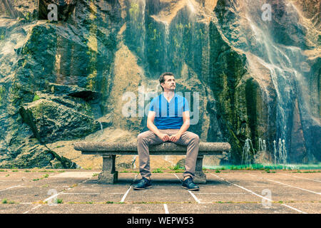 man sitting on a bench next to waterfall thinking and looking up Stock Photo