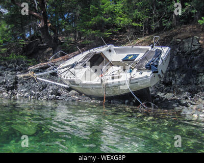Sunlight reflects off the water onto the hull of a wrecked fibreglass sailboat, heeled over and hard aground on a rocky beach in British Columbia. Stock Photo