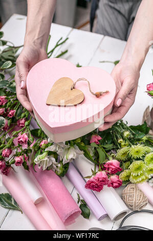 Female hands holding a gift box in the shape of a heart. Florist's workplace: flowers, accessories, tools. Stock Photo