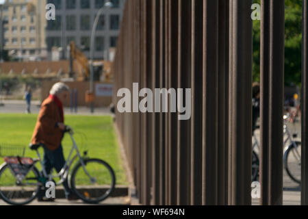 Elder berliner cyclist woman dressed in a brown coat near a grass field looking at the metal bars of the Memorial of the Berlin Wall at the Memorial P Stock Photo