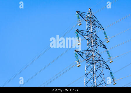 View from below of an electricity pylon supporting an overhead high-voltage power line against blue sky. Stock Photo