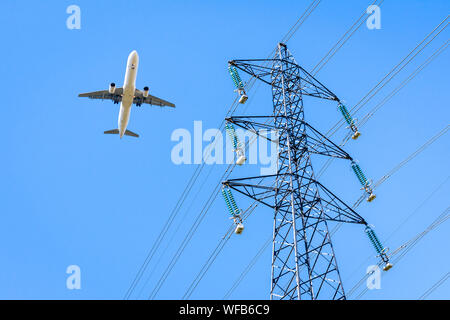 View from below of an airliner in landing approach flying over a high-voltage power line against blue sky with an electricity pylon in the foreground. Stock Photo