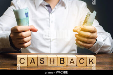 Wooden blocks with the word Cashback and money in the hands of a businessman. Bonus program to attract customers and increase their loyalty. Internet Stock Photo