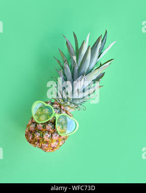 Funny pineapple in heart-shaped sunglasses, flat lay on neo green paper background Stock Photo