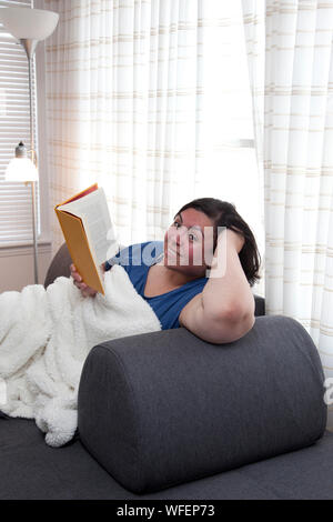 Happy hispanic woman is reading a book on her sofa, smiling and content Stock Photo