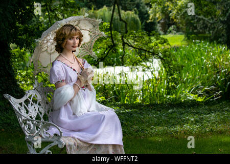 Beautiful lady in vintage dress sitting on ornate metal bench holding parasol next to river Stock Photo