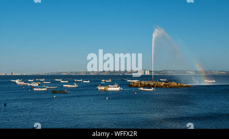 Geyser forming a rainbow on River Tagus with fishing boats overlooking 25 April Bridge and Rei Cristo statue in Lisbon, Portugal with copy space Stock Photo