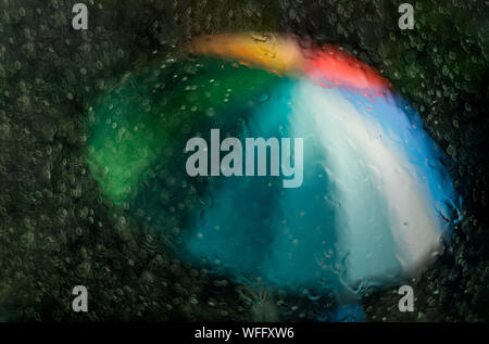 A multicolored rainbow umbrella with red, orange, yellow, green, blue, and purple is seen through a window that is wet with rain at nighttime Stock Photo