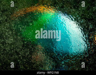 A multicolored rainbow umbrella with red, orange, yellow, green, blue, and purple is seen through a window that is wet with rain at nighttime Stock Photo