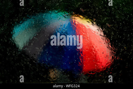 A multicolored rainbow umbrella with red, orange, yellow, green, blue, and purple is seen through a window that is wet with rain at nighttime Stock Photo