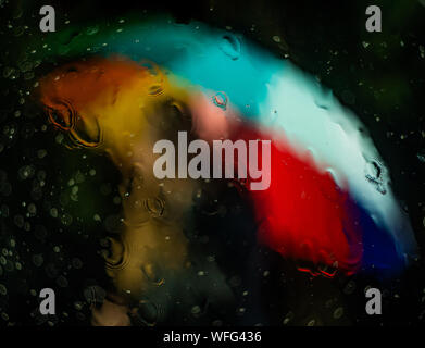 A multicolored rainbow umbrella with red, orange, yellow, green, blue, and purple is seen through a window that is wet with rain at nighttime Stock Photo