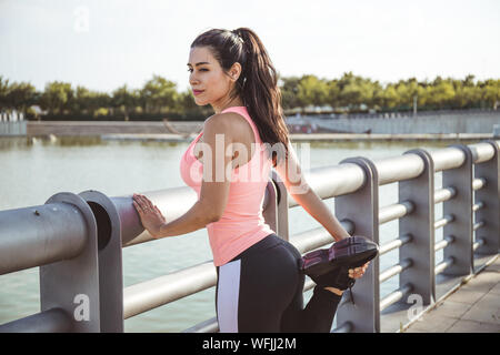 Latin woman does muscle stretching in front of a lake and dressed in pink top and black pants Stock Photo