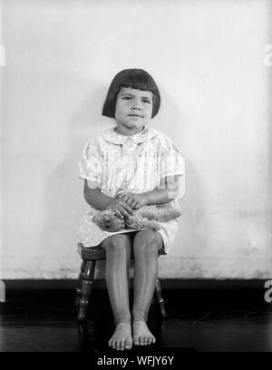 A vintage late Victorian or early Edwardian black and white photograph showing a young girl with a short bobbed haircut, sitting in a chair facing the camera. She is barefoot, and holding a toy teddy bear. Stock Photo