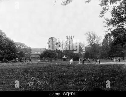 A vintage late Victorian or early Edwardian black and white photograph showing a game of cricket being played in an English park or garden, lithesome people watching on.