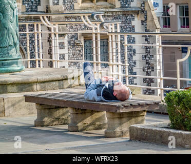 Norwich, Norfolk, UK – August 31 2019. Young male sleeping off a hangover from the night before on a concrete bench in a city park Stock Photo