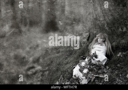 A ceepy spooky doll, sitting in a forest in winter, next to a tree. With a textured, weathered, sepia edit. Stock Photo