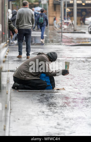 A homeless person kneeling holding a McDonalds cup out and begging on a wet and rainy footpath in the Sydney CBD Stock Photo