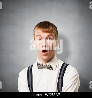 Stupefied teenager with mouth opened. Emotional redhead boy has shocked facial expression. Portrait of guy wears white shirt, bow tie and suspenders o Stock Photo