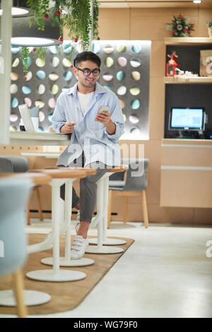Full length portrait of mixed race college student smiling while looking at smartphone screen in cafe, copy space Stock Photo