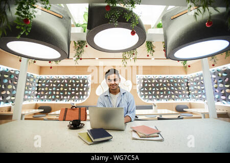 Wide angle portrait of Middle-Eastern young man using laptop while studying in cafe and smiling at camera, copy space Stock Photo