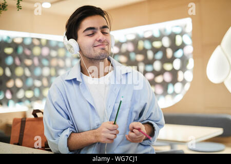 Waist up portrait of smiling Middle-Eastern man wearing headphones listening to music and air drumming beat with pencils, copy space Stock Photo