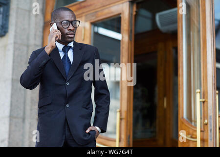 Waist up portrait of stylish African-American man wearing suit speaking by smartphone while leaving work, copy space Stock Photo